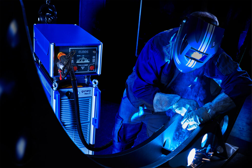 A welder in protective gear manually welds a curved steel part, sparks visible, with a CLOOS QINEO NexT power source and live weld parameters shown in the foreground.