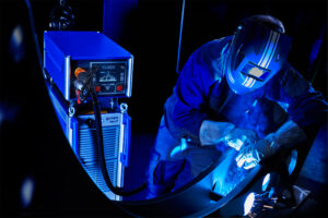 A welder in protective gear manually welds a curved steel part, sparks visible, with a CLOOS QINEO NexT power source and live weld parameters shown in the foreground.