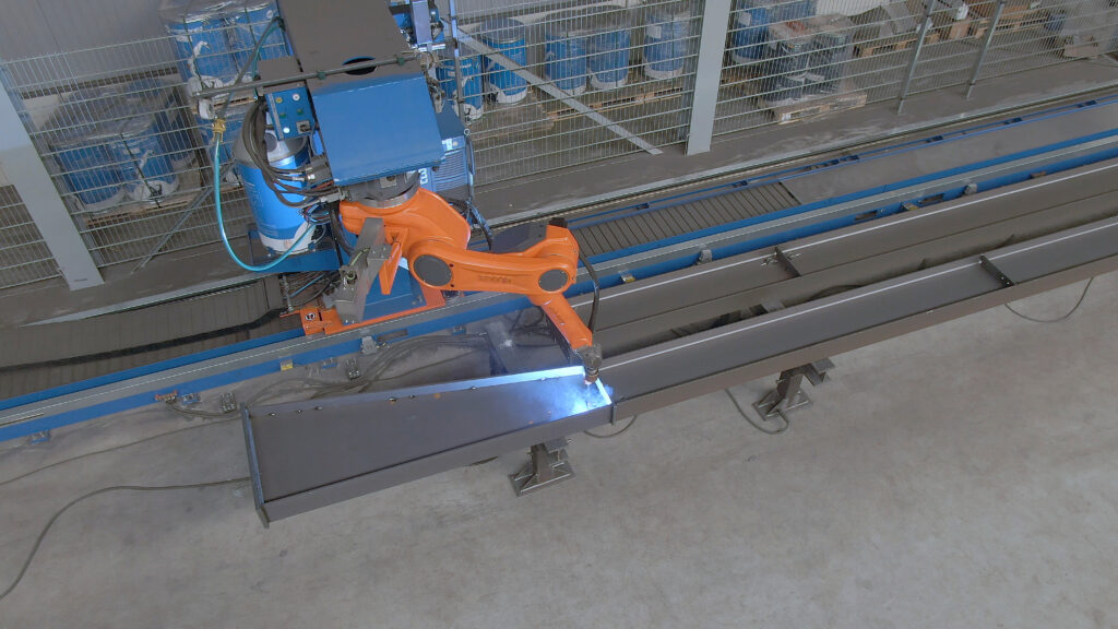 Orange CLOOS robot arm on a rail-mounted carriage welding a long steel beam, with a bright blue arc at the torch.